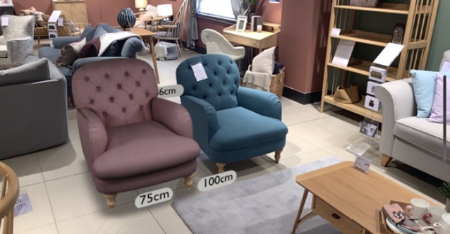 A shop floor: a virtual teal armchair with 75cm and 100cm dimension labels attached, placed beside real armchairs and furniture in a John Lewis showroom.