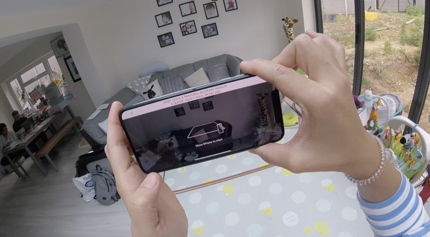 Head-mounted camera view: a participant holds an iPhone running the sofa AR feature in their living room, their existing sofa and family photographs behind it.